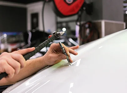 Mains réparant une bosse sur une carrosserie de voiture blanche avec des outils à Brissac Quince dans le Maine-et-Loire 49